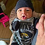 baby, infant, clothing, hat, person, indoor, holding, hand, face, expression, curious, black_hat, jaguars, football, pacifier_clip, wooden_floor, pink_chair, plant, tattoo, casual