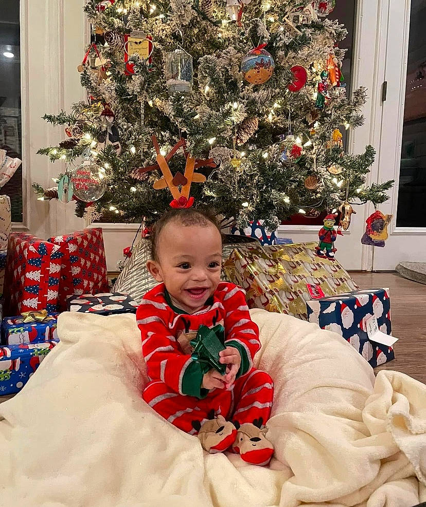 Judah is registered to the contest to win money with this photo: beauty, christmas_decoration, christmas_ornament, christmas_tree, comfort, event, evergreen, facial_expression, happy, holiday_ornament, human_body, lap, leaf, living_room, ornament, person, plant, red, smile, toddler