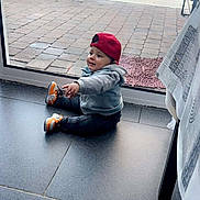 Zayn a rejoint le concours — aidez-le/la à gagner de superbes lots ! toddler, child, indoor, casual_clothing, red_cap, gray_hoodie, sneakers, sitting, curious, glass_door, backyard, garden, paved_floor, tiles, daylight, home, person, young_child, floor, window