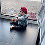 toddler, child, indoor, casual_clothing, red_cap, gray_hoodie, sneakers, sitting, curious, glass_door, backyard, garden, paved_floor, tiles, daylight, home, person, young_child, floor, window