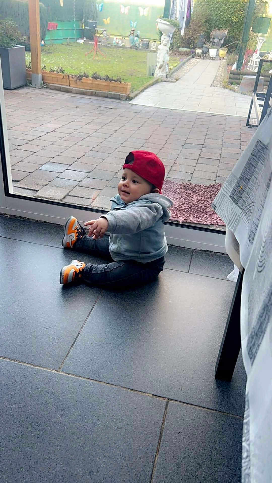 Zayn a rejoint le concours — aidez-le/la à gagner de superbes lots ! toddler, child, indoor, casual_clothing, red_cap, gray_hoodie, sneakers, sitting, curious, glass_door, backyard, garden, paved_floor, tiles, daylight, home, person, young_child, floor, window