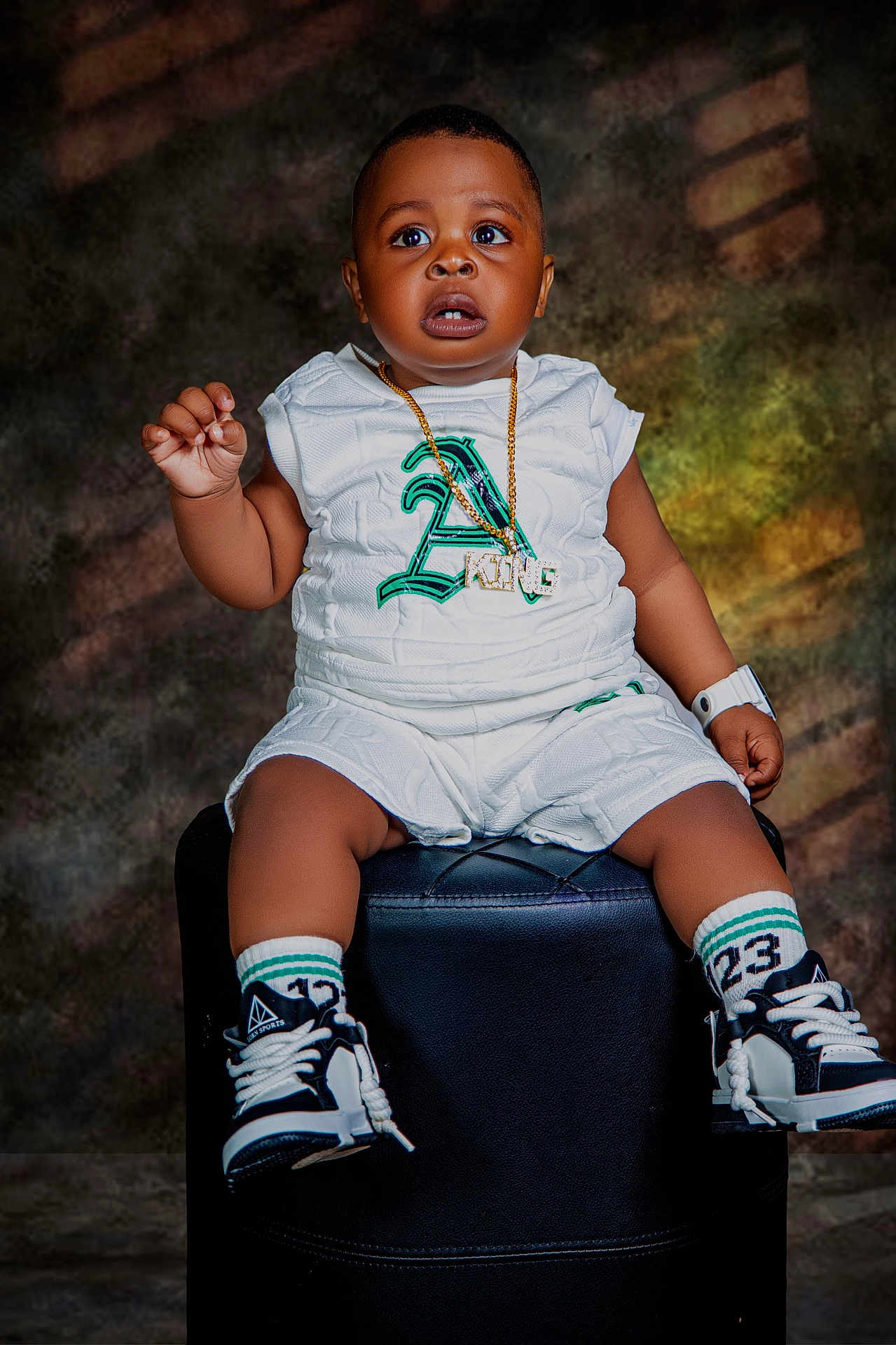 David is registered to the contest to win money with this photo: toddler, child, sitting, white_clothing, sneakers, gold_chain, watch, black_ottoman, curious_expression, studio_lighting, portrait, shorts, socks, fashion, cute, indoor, person, footwear, accessories, young_child