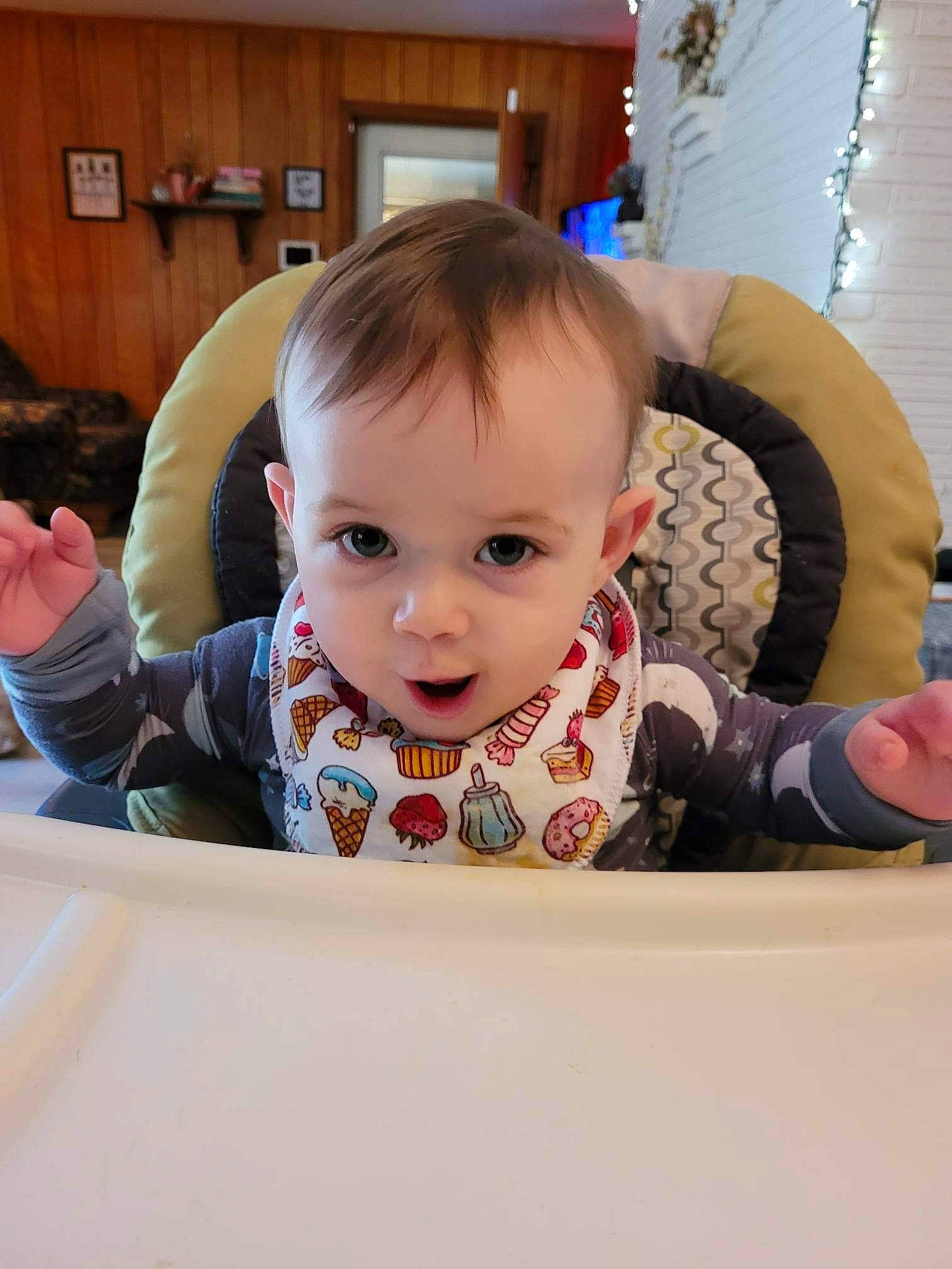 Solomon is registered to the contest to win money with this photo: baby, baby_toddler_clothing, cabinetry, chair, cheek, child, floor, flooring, fun, happy, person, play, room, sitting, sleeve, smile, surprise, t_shirt, table, toddler