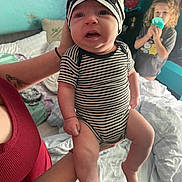 Jackson is registered to the contest to win money with this photo: baby, infant, child, toddler, person, striped_clothing, hat, holding, bedroom, tattoo, adult_hand, curly_hair, cup, blanket, wall, furniture, indoor, expression, wearing, sitting
