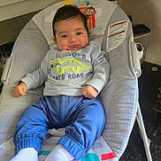 Kylian is registered to the contest to win money with this photo: baby, child, infant, portrait, smiling, sitting, bouncer, baby_seat, dinosaur, sweatshirt, blue_pants, white_socks, cartoon, monkey, indoor, home, kitchen, furniture, cozy, happy