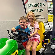 Ava is registered to the contest to win money with this photo: child, boy, girl, tractor, indoor, john_deere, steering_wheel, smile, yellow_seat, banner, sign, clothing, shorts, tshirt, red_hair, happy, toy, vehicle, play, fun