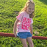 child, girl, smiling, pink_tshirt, bow, hair, grass, outdoor, sunlight, shorts, red_bar, summer, playground, happy, casual_clothing, nature, young, person, sitting, daytime