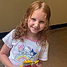 Ava is registered to the contest to win money with this photo: child, girl, smile, red_hair, curly_hair, tshirt, cartoon_characters, rainbow, snack_bags, indoor, floor, wall, person, portrait, happy, holding, cute, casual_clothing, young, light