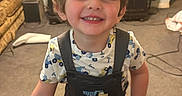 Asher is registered to the contest to win money with this photo: cabinet, carpet, carpeted_floor, casual_clothing, child, couch, cute, face, furniture, hair, happy, home, indoor, living_room, overalls, person, smiling, toddler, toy_pattern, young_child
