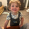child, smiling, indoor, toddler, overalls, carpet, furniture, couch, toy_pattern, hair, face, happy, living_room, cabinet, carpeted_floor, person, casual_clothing, young_child, cute, home