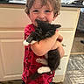 child, kitten, pet, hug, smile, curly_hair, red_shirt, indoor, kitchen, cabinet, floor, happy, person, animal, feline, cute, domestic, young, black_and_white, casual