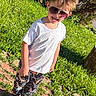 child, boy, sunglasses, white_tshirt, colorful_pants, dinosaur_print, outdoor, grass, sunlight, smiling, standing, shoes, casual_clothing, nature, greenery, playful, young_child, happy, daylight, tree