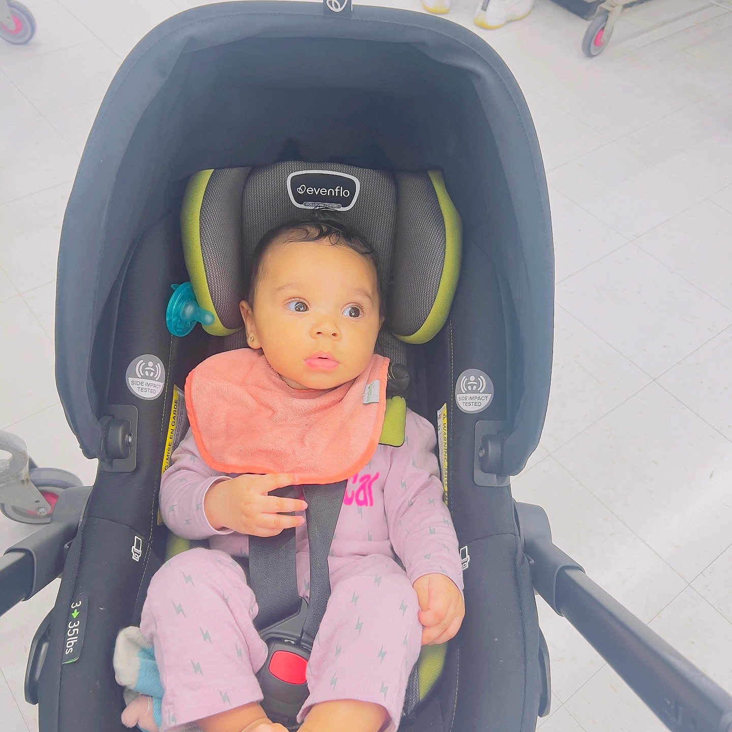Zula joined the competition — help win amazing prizes! baby, bare_feet, bib, bright_lighting, car_seat, child, curly_hair, evenflo, floor_tiles, footwear_absent, hands, indoor, infant, looking_away, pacifier, person, pink_clothing, seatbelt, sitting, stroller_handle