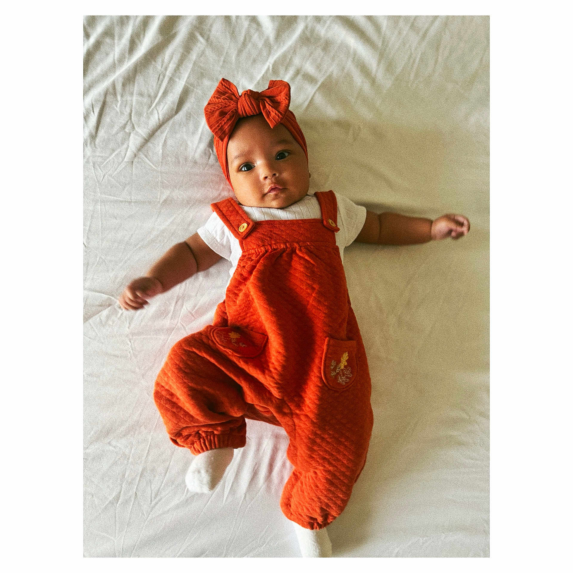 Kamila joined the competition — help win amazing prizes! baby, infant, child, orange_clothing, headband, bow, white_shirt, lying_down, bed, sheet, cute, portrait, face, hands, feet, one_person, indoors, soft_lighting, textured_fabric, expression