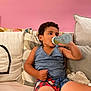 bottle, casual_clothing, cat_illustration, child, colorful, comfort, couch, cozy, curly_hair, drinking, home, indoor, patterned, pillows, playful, plush_toy, relaxed, seated, smiling, young_child