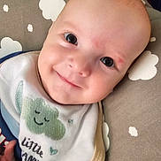 Zayden is registered to the contest to win money with this photo: baby, smiling, bib, blanket, cushion, cloud_pattern, infant, face, child, happy, portrait, indoor, cute, clothing, head, person, soft_toys, comfort, cozy, resting