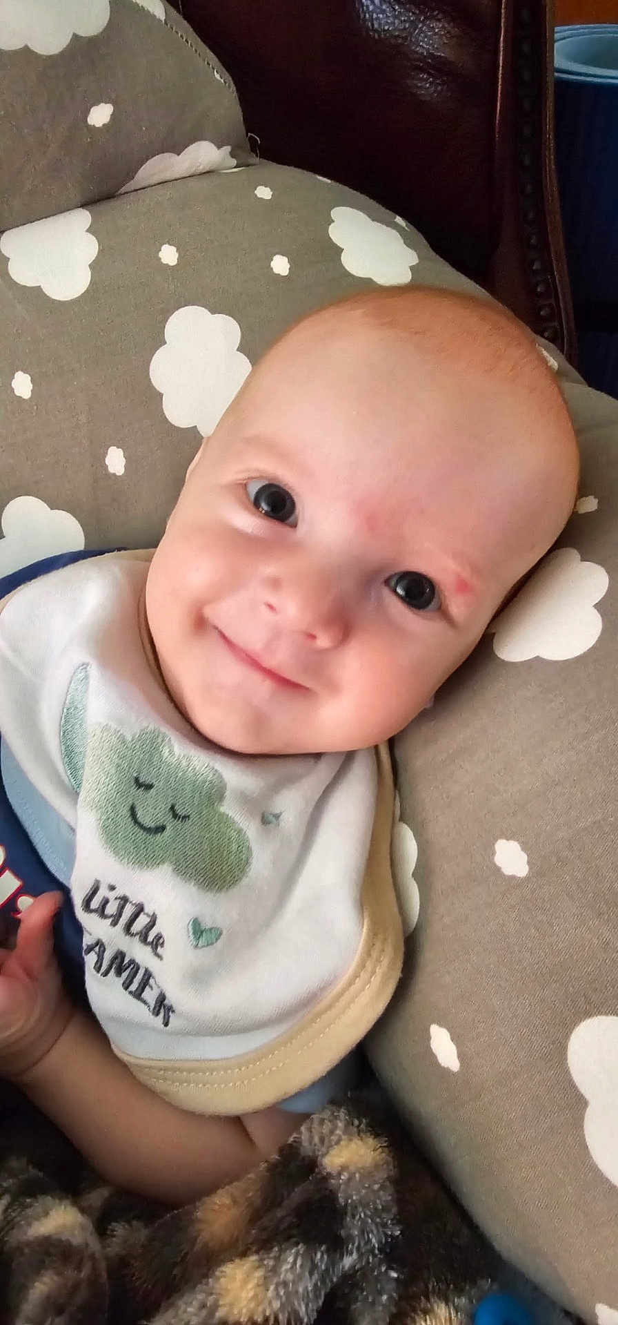 Zayden is registered to the contest to win money with this photo: baby, smiling, bib, blanket, cushion, cloud_pattern, infant, face, child, happy, portrait, indoor, cute, clothing, head, person, soft_toys, comfort, cozy, resting