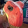 dog, dinosaur_mask, red_mask, pet, costume, eye, hand, indoor, cluttered_background, toy, animal, face_covering, closeup, texture, scales, mouth, teeth, floor, person_hand, funny