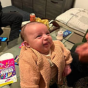 Nahila a rejoint le concours — aidez-le/la à gagner de superbes lots ! baby, infant, knitted_cardigan, floral_pants, funny_face, indoor, coffee_table, can, smartphone, bottle, couch, pillow, person, seated, carpet, hand, clothing, furniture, expression, home