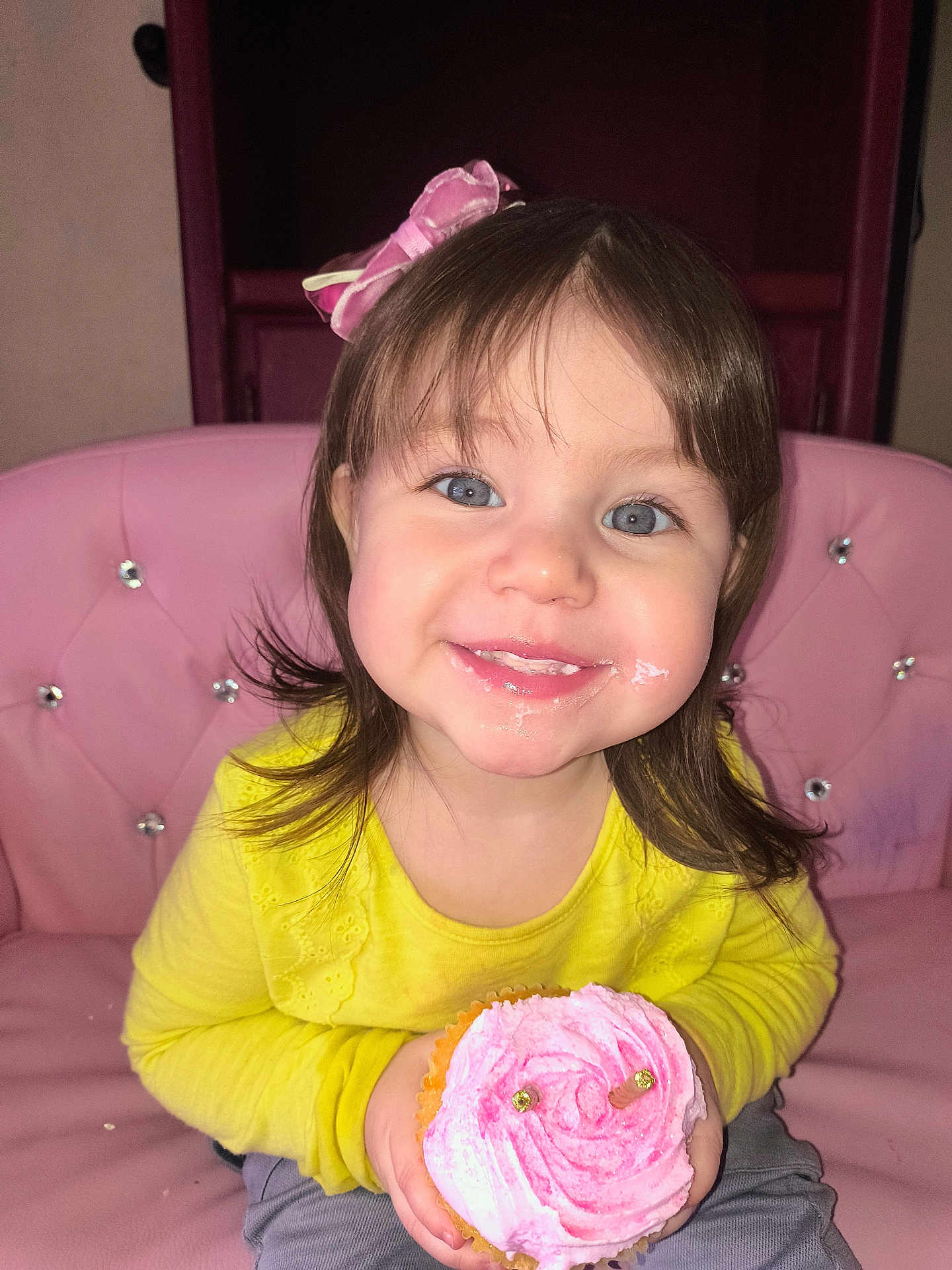 Dulce Sofia is registered to the contest to win money with this photo: blue_eyes, celebration, child, cupcake, cute, face, food, hair, happy, indoors, pink_bow, pink_chair, pink_frosting, portrait, sitting, small_hands, smile, sweet, toddler, yellow_shirt