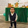 balloon, birthday_party, celebration, child, curious_expression, decorations, floor, green_tablecloth, indoor, lion_king, pants, party, person, shoes, smiling, table, toddler, vest, wooden_floor, young_child