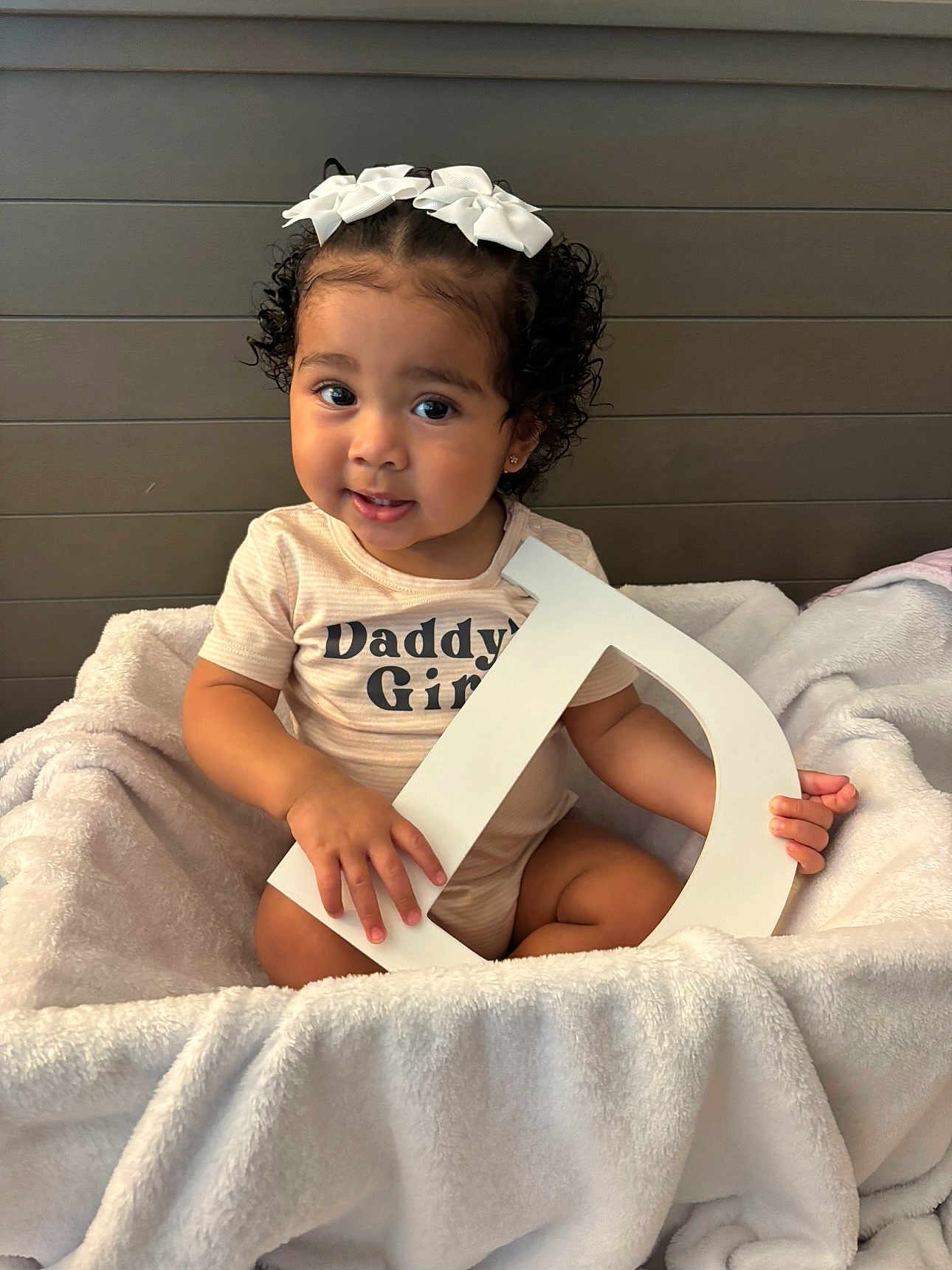 Arabella is registered to the contest to win money with this photo: toddler, child, girl, curly_hair, white_bows, blanket, letter_d, shirt, daddys_girl, sitting, indoor, portrait, cute, smiling, baby, soft_texture, cozy, person, furniture, wall