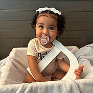 Arabella is registered to the contest to win money with this photo: baby, pacifier, blanket, basket, curly_hair, bow, letter, wooden_headboard, indoor, cute, child, smiling, seated, soft_texture, infant, holding, light_skin, portrait, cozy, home
