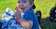 Caysen is registered to the contest to win money with this photo: baby, child, toddler, smiling, happy, outdoor, portrait, tennis_court, sports, tennis, grass, blanket, blue_shirt, cap, sitting, stroller, sunlight, fence, flag, family_event