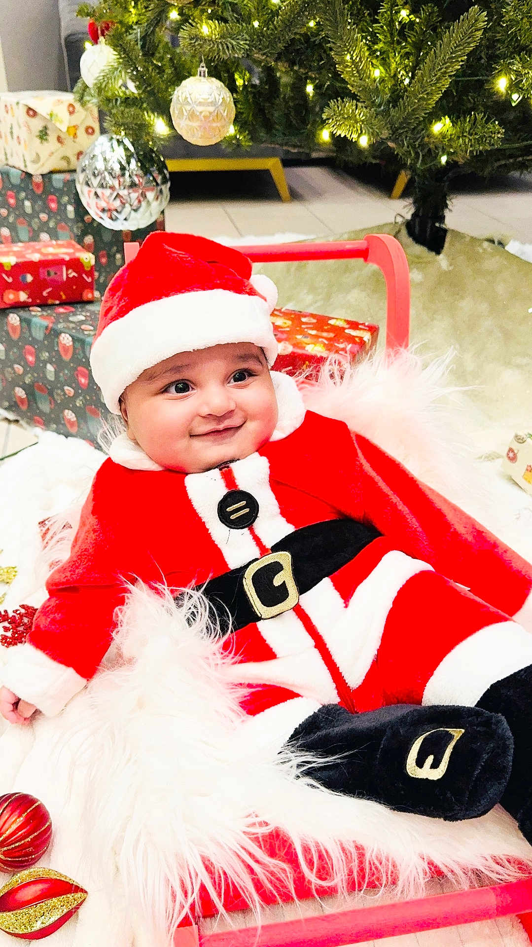 Shivin joined the competition — help win amazing prizes! baby, black_belt, blanket, child, christmas_ornaments, christmas_tree, cute, decorations, face, festive, floor_tiles, holiday, holiday_season, indoors, portrait, presents, red_clothing, santa_claus_costume, smiling, white_fur