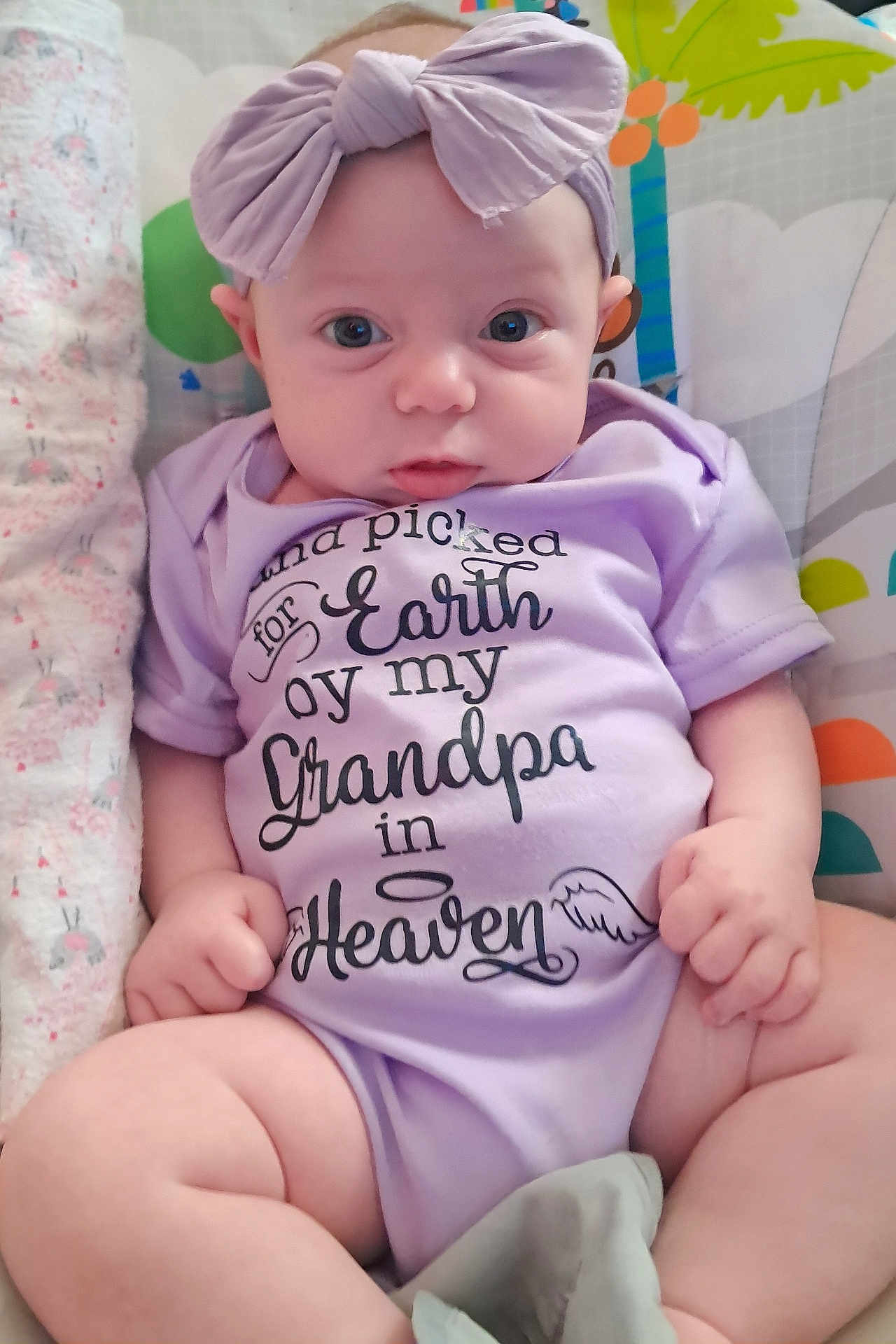 Leanna Mae joined the competition — help win amazing prizes! baby, infant, onesie, headband, bow, purple, eyes, face, hands, legs, blanket, pattern, text_on_clothing, sitting, portrait, close_up, cute, indoor, soft_toys, nursery