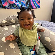 Mezekiah is registered to the contest to win money with this photo: baby, infant, smiling, happy, sitting, clothing, green_bib, green_pants, white_sock, indoor, pillow, cloud_pattern, face, person, hair_buns, tapestry, colorful, background, pacifier, bed
