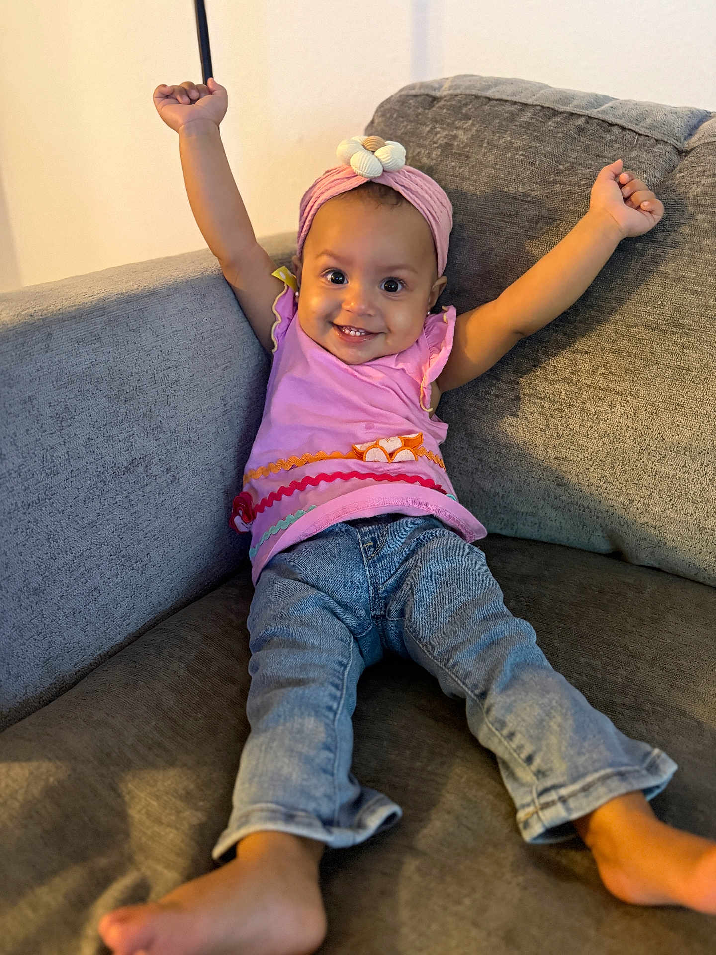 Aria is registered to the contest to win money with this photo: baby, child, smile, happy, couch, jeans, headband, pink, arms_up, indoor, person, clothing, barefoot, cute, infant, sitting, furniture, portrait, joyful, casual
