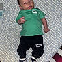 baby, infant, child, green_shirt, black_pants, all_star, footwear, blanket, pattern, bed, hand, face, eyes, expression, indoor, cute, clothing, person, lying_down, cozy