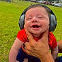 baby, headphones, smile, hand, outdoor, grass, person, child, happy, red_shirt, blue_pants, socks, park, nature, trees, blurred_background, holding, cute, infant, joy