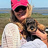 woman, dog, puppy, cap, pink_cap, sunglasses, smile, outdoor, nature, grass, water, sky, striped_cloth, holding, portrait, happy, animal, cute, casual_clothing, daylight