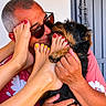 man, dog, feet, toenails, red_nail_polish, yellow_nail_polish, sunglasses, hug, affection, smile, short_hair, casual_clothing, shirt, hand, pet, indoor, skin, close_up, playful, funny