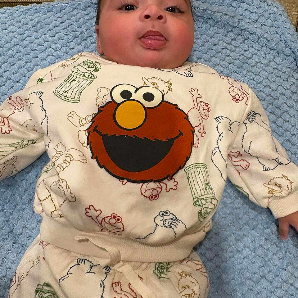 Makari is registered to the contest to win money with this photo: baby, blue_blanket, child, clothing, cozy, cute, elmo, face, indoors, infant, lying_down, patterned_clothing, playful, portrait, sesame_street, sleepsuit, smiling, soft_texture, tongue, young_child