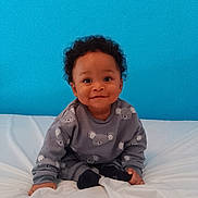 Joari a rejoint le concours — aidez-le/la à gagner de superbes lots ! baby, child, curly_hair, smiling, sitting, bed, blue_wall, koala_pattern, sweatshirt, socks, indoor, cute, portrait, happy, young_child, person, infant, cozy, casual_clothing, bright_background