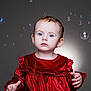 child, toddler, baby, blue_eyes, red_dress, velvet, bubbles, studio_lighting, portrait, curious, hands, soft_light, indoor, innocent, cute, fashion, young_child, face, expression, studio_background