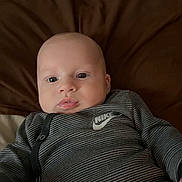 Draven is registered to the contest to win money with this photo: baby, infant, face, onesie, nike, striped_clothing, blanket, brown_blanket, young_child, portrait, cute, lying_down, indoors, child, person, apparel, comfortable, expression, looking_at_camera, skin