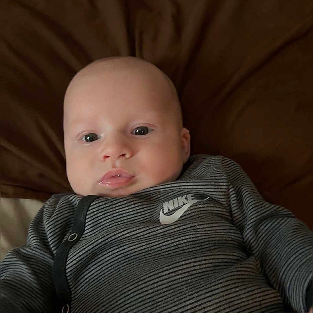 Draven is registered to the contest to win money with this photo: apparel, baby, blanket, brown_blanket, child, comfortable, cute, expression, face, indoors, infant, looking_at_camera, lying_down, nike, onesie, person, portrait, skin, striped_clothing, young_child