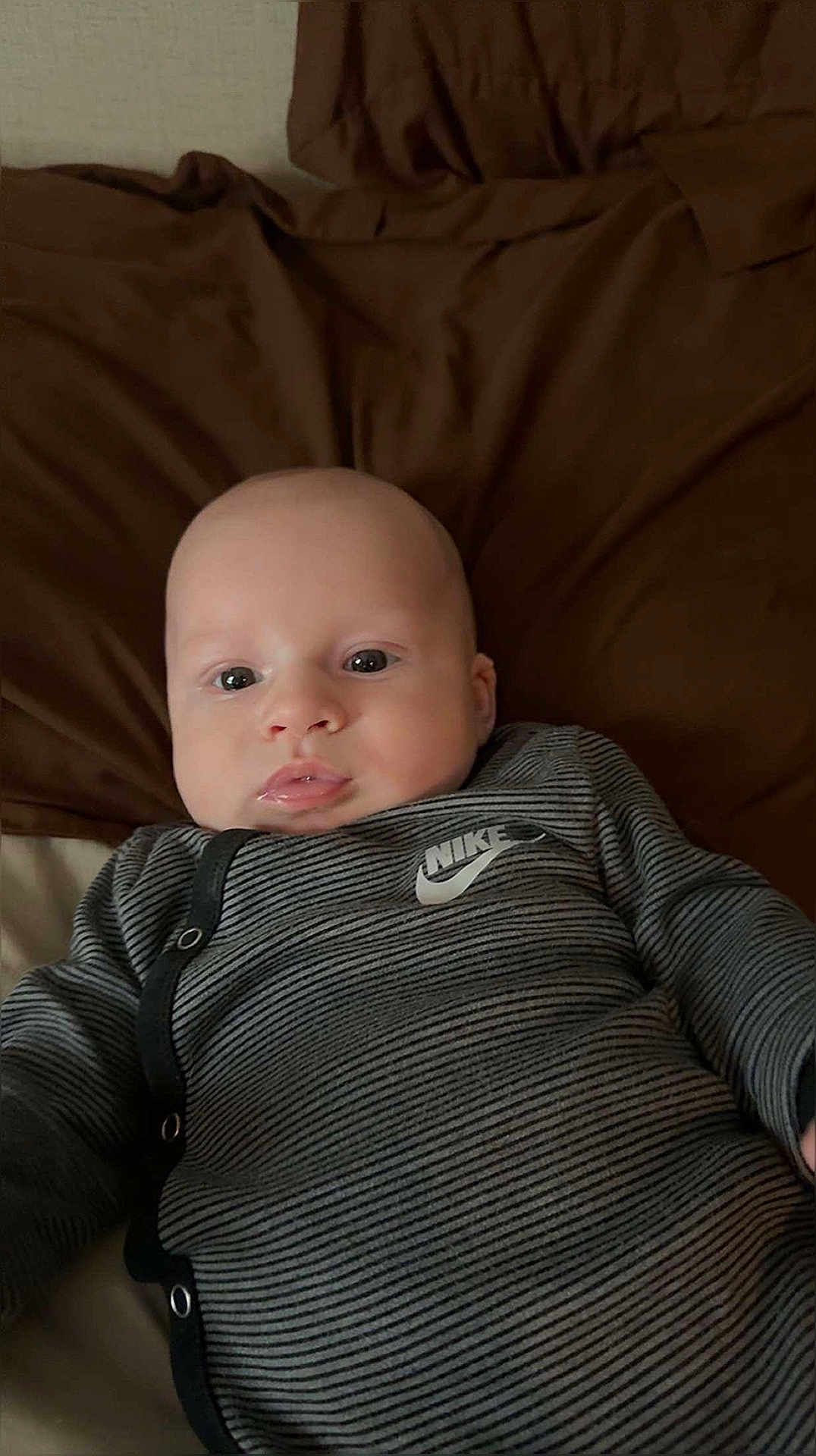 Draven is registered to the contest to win money with this photo: baby, infant, face, onesie, nike, striped_clothing, blanket, brown_blanket, young_child, portrait, cute, lying_down, indoors, child, person, apparel, comfortable, expression, looking_at_camera, skin