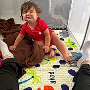 Adonis joined the competition — help win amazing prizes! toddler, child, red_onesie, playpen, colorful_mat, dinosaur_pattern, blanket, brown_blanket, happy, smiling, sitting, indoor, person, legs, socks, hair, cute, baby, fun, playtime