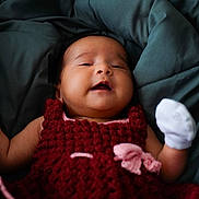 Ira is registered to the contest to win money with this photo: baby, infant, smiling, crocheted_dress, red_clothing, white_mitten, blanket, green_blanket, cozy, closeup, portrait, child, cute, happy, lying_down, soft_texture, indoors, person, newborn, warm