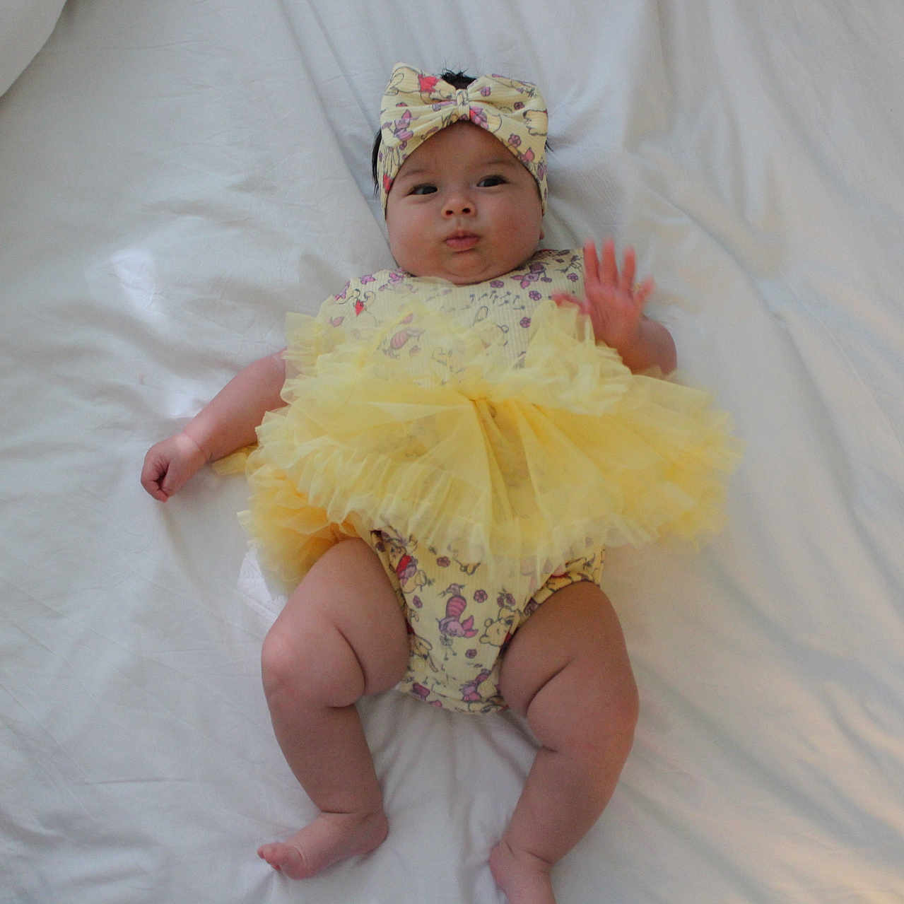 Lillyann is registered to the contest to win money with this photo: baby, bed, bedding, child, chubby, clothing, cute, hand, headband, indoors, infant, legs, lying_down, person, smile, soft, tutu, waving, white, yellow