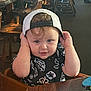 child, toddler, cap, backwards_cap, black_shirt, patterned_shirt, high_chair, wooden_chair, indoor, restaurant, curly_hair, blue_eyes, hands, face, table, wooden_table, cute, portrait, person, baby
