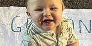 Greyson is registered to the contest to win money with this photo: toddler, child, smiling, curly_hair, shirt, dinosaur_print, towel, carpet, playmat, baby, cute, sitting, happy, indoors, portrait, face, young_child, clothing, infant, teeth
