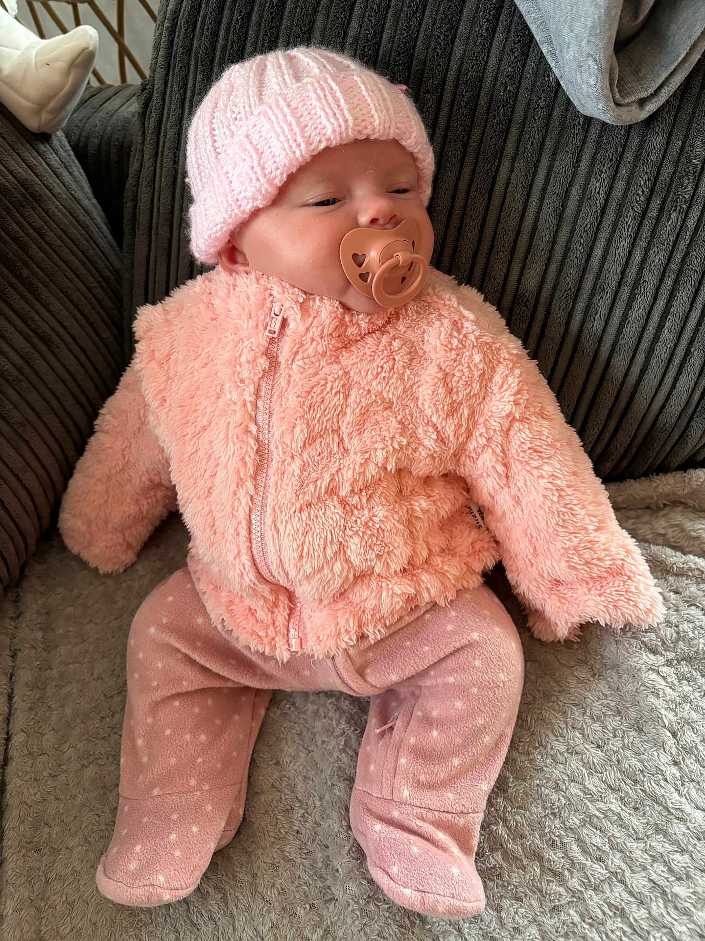 Marnie is registered to the contest to win money with this photo: baby, pacifier, pink_clothing, knitted_hat, fluffy_jacket, infant, couch, blanket, polka_dot_pants, child, indoor, soft_texture, seated, cute, cozy, warm_clothing, face, person, relaxing, home