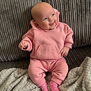 Marnie is registered to the contest to win money with this photo: baby, child, pink_hoodie, pink_pants, pink_socks, smiling, happy, couch, blanket, indoors, cozy, seated, infant, face, clothing, person, cute, soft_texture, home, relaxed