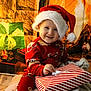 toddler, child, santa_hat, red_pajamas, christmas_present, gift_wrapping, holiday_decor, fireplace, christmas_lights, smiling, indoors, festive, cozy, warm_lighting, holiday_season, wrapped_gifts, floor, blanket, happy, portrait